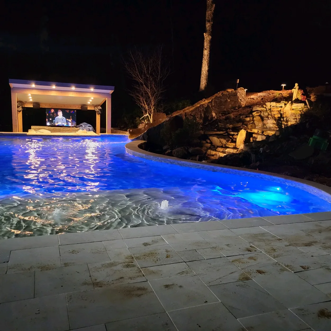 Pool glowing blue at night next to a lit cabana and stone waterfall